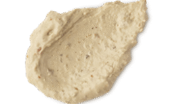 A medium circular puddle of some creamy beige face mask that has some pieces of dried fig running through it, on a white background.