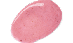 A medium circular puddle of some creamy pastel pink body scrub that has some pieces of Tagua Nut Powder, Ground White Rice, and Pomegranate Seed Powder running through it, on a white background.,
