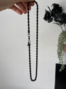 A hand holding a metallic black rope with a metallic silver clasp and a small metal charm that has a Letter E engraved on it, on a white background.