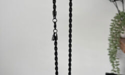 A hand holding a metallic black rope with a metallic silver clasp and a small metal charm that has a Letter E engraved on it, on a white background.