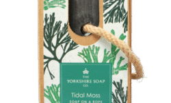 A medium rectangular dark blue block of soap inside a medium rectangular cardboard box with a dark green label that has The Yorkshire Soap Company Tidal Moss Soap On A Rope Bar written in bold bright white writing, some pictures of some bright green pieces of Seaweed, and a piece of light brown rope tied around it, on a white background.