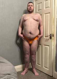 A tall bearded man with his hands on his hips wearing a bright orange brief with a bright orange waistband that has Kiniki written in bright white writing on it stood against a white wooden door.