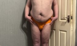 A tall bearded man with his hands on his hips wearing a bright orange brief with a bright orange waistband that has Kiniki written in bright white writing on it stood against a white wooden door.
