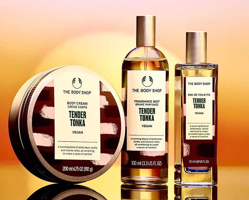 The Tender Scent Of Tonka Is So Sensual And Alluring | The Body Shop Tender Tonka Range A short cylindrical dark green plastic tub full of some creamy white body cream with a bright white label that has The Body Shop Tender Tonka Body Cream written in dark green writing and a dark brown and pink brushstroke pattern on it next to a tall cylindrical clear glass bottle with a silver metallic lid and a bright white label that has The Body Shop Tender Tonka Fragrance Mist written in dark green writing and a dark brown and pink brushstroke pattern on it, and a tall rectangular glass bottle with a silver metallic lid and a bright white label that has The Body Shop Tender Tonka Eau De Toilette written in dark green writing and a dark brown and pink brushstroke pattern on it on a light wooden shelf, on a white background.