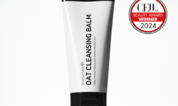 A tall conical bright white plastic tube with a black plastic lid that has The INKEY List Oat Cleansing Balm written in bold black writing on it, on a white background.