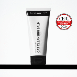 A tall conical bright white plastic tube with a black plastic lid that has The INKEY List Oat Cleansing Balm written in bold black writing on it, on a white background.