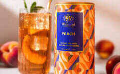 A tall cylindrical gradient bright blue, orange and peach geometric patterned cardboard tube with a dark blue plastic lid and a dark blue label that has Whittard of Chelsea peach Instant Tea written in bright gold writing on it next to a tall cylindrical clear glass full of some translucent amber liquid and some pieces of bright pink peaches with a bright white straw coming out of the top of it, on a white background.