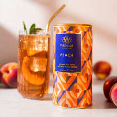 Tea Tastes Terrific | Whittard Of Chelsea Peach & Pomelo Instant Teas A tall cylindrical gradient bright blue, orange and peach geometric patterned cardboard tube with a dark blue plastic lid and a dark blue label that has Whittard of Chelsea peach Instant Tea written in bright gold writing on it next to a tall cylindrical clear glass full of some translucent amber liquid and some pieces of bright pink peaches with a bright white straw coming out of the top of it, on a white background.