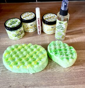 A small oval bright green soap sponge wrapped in some clear plastic next to a large oval bright green soap sponge wrapped in some clear plastic, a short cylindrical bright white plastic tub that has Bath Bomb Bliss Mojito Whipped Soap written in bold black writing and some pictures of some slices of lime on it, a short cylindrical bright white plastic tub that has Bath Bomb Bliss Pineapple Whipped Soap written in bold black writing and some pictures of some pineapples on it, a short cylindrical bright white plastic tub that has Bath Bomb Bliss pineapple body scrub written in bold black writing and some pictures of some pineapples on it, a tall cylindrical bright white plastic spray bottle that has Bath Bomb Bliss Mojito Body Mist written in bold black writing and some pictures of some slices of lime on it, and a small cylindrical bright white plastic spray bottle that has Bath Bomb Bliss Cherry Body mist written in bold black writing on it on a light wooden worktop.