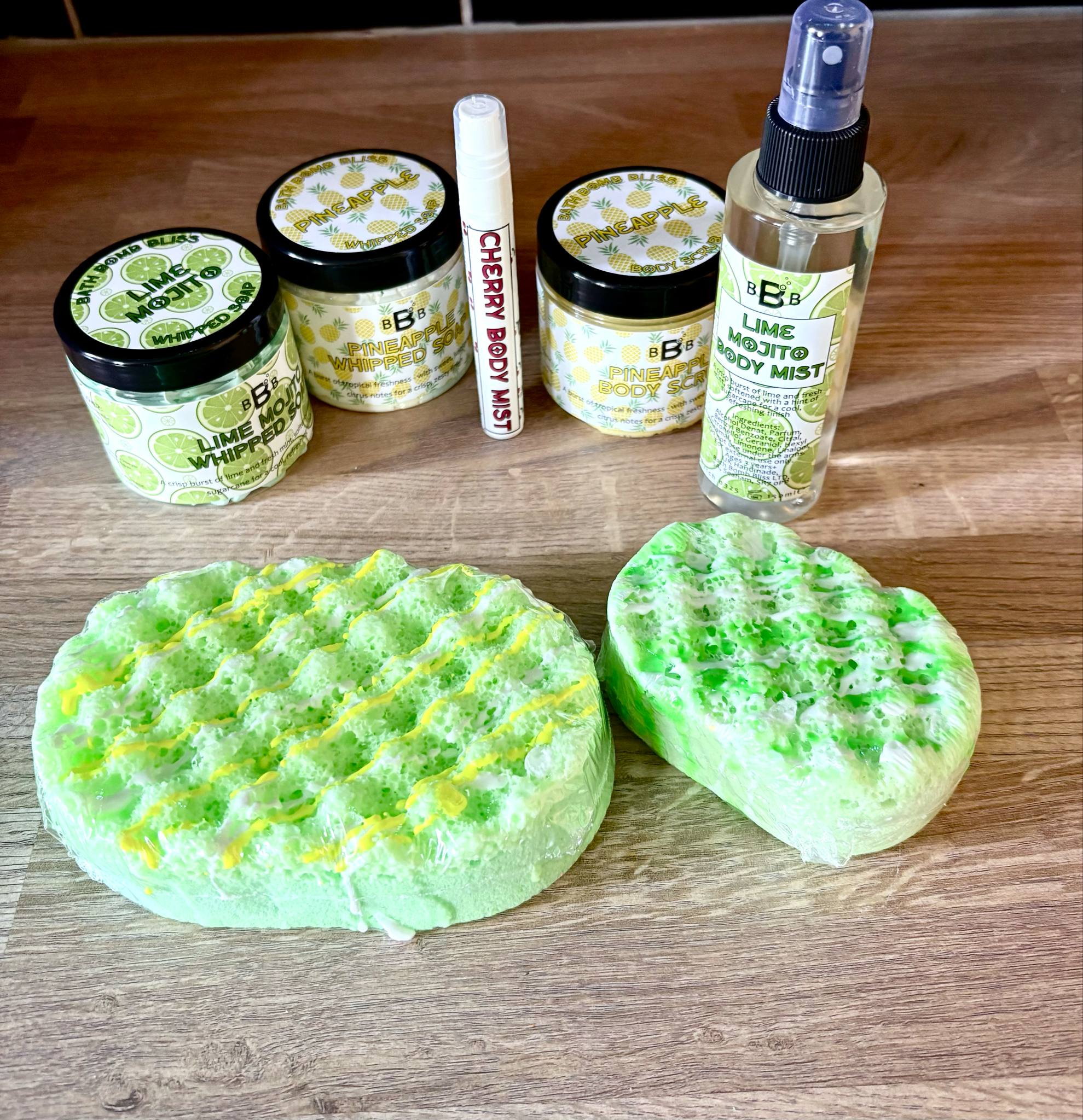 A small oval bright green soap sponge wrapped in some clear plastic next to a large oval bright green soap sponge wrapped in some clear plastic, a short cylindrical bright white plastic tub that has Bath Bomb Bliss Mojito Whipped Soap written in bold black writing and some pictures of some slices of lime on it, a short cylindrical bright white plastic tub that has Bath Bomb Bliss Pineapple Whipped Soap written in bold black writing and some pictures of some pineapples on it, a short cylindrical bright white plastic tub that has Bath Bomb Bliss pineapple body scrub written in bold black writing and some pictures of some pineapples on it, a tall cylindrical bright white plastic spray bottle that has Bath Bomb Bliss Mojito Body Mist written in bold black writing and some pictures of some slices of lime on it, and a small cylindrical bright white plastic spray bottle that has Bath Bomb Bliss Cherry Body mist written in bold black writing on it on a light wooden worktop.