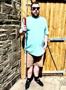 A tall bearded man wearing a pair of shiny denim blue sunglasses with some bright red lenses, a bright turquoise crew net t-shirt, a pair of stone grey shorts and a pair of black trainers with some white souls holding a bright white cane with a red leather handle stood against a light wooden fence.