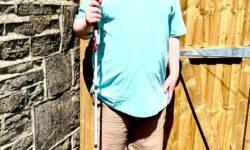A tall bearded man wearing a pair of shiny denim blue sunglasses with some bright red lenses, a bright turquoise crew net t-shirt, a pair of stone grey shorts and a pair of black trainers with some white souls holding a bright white cane with a red leather handle stood against a light wooden fence.