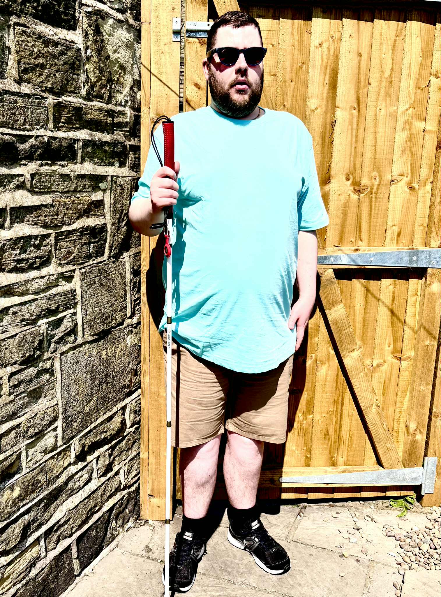 A tall bearded man wearing a pair of shiny denim blue sunglasses with some bright red lenses, a bright turquoise crew net t-shirt, a pair of stone grey shorts and a pair of black trainers with some white souls holding a bright white cane with a red leather handle stood against a light wooden fence.