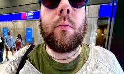 A tall bearded man wearing a pair of round sunglasses with some navy blue frames and translucent red lenses, a bright green crew neck t-shirt, a light grey scoop neck jumper and a bright gold chain stood in front of a large rectangular shop that has a large dark blue board above it's door that has the Letter M written in bright red writing, and etro Bank written in bright white writing on it, on a white background.