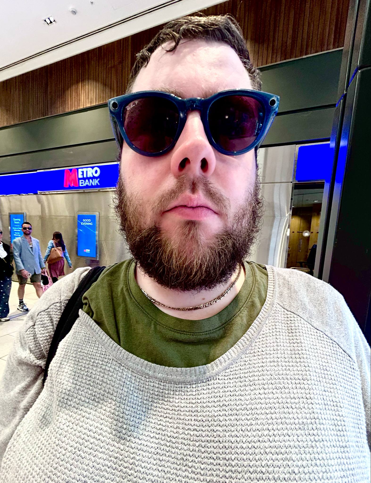 A tall bearded man wearing a pair of round sunglasses with some navy blue frames and translucent red lenses, a bright green crew neck t-shirt, a light grey scoop neck jumper and a bright gold chain stood in front of a large rectangular shop that has a large dark blue board above it's door that has the Letter M written in bright red writing, and etro Bank written in bright white writing on it, on a white background.