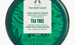 A short cylindrical dark green plastic tub with a dark green plastic lid and a bright green label that has The Body Shop Tea tree Purifying & Balancing Hair & Scalp Scrub written in dark green writing and some pictures of some dark green tea tree leaves on it, on a white background.