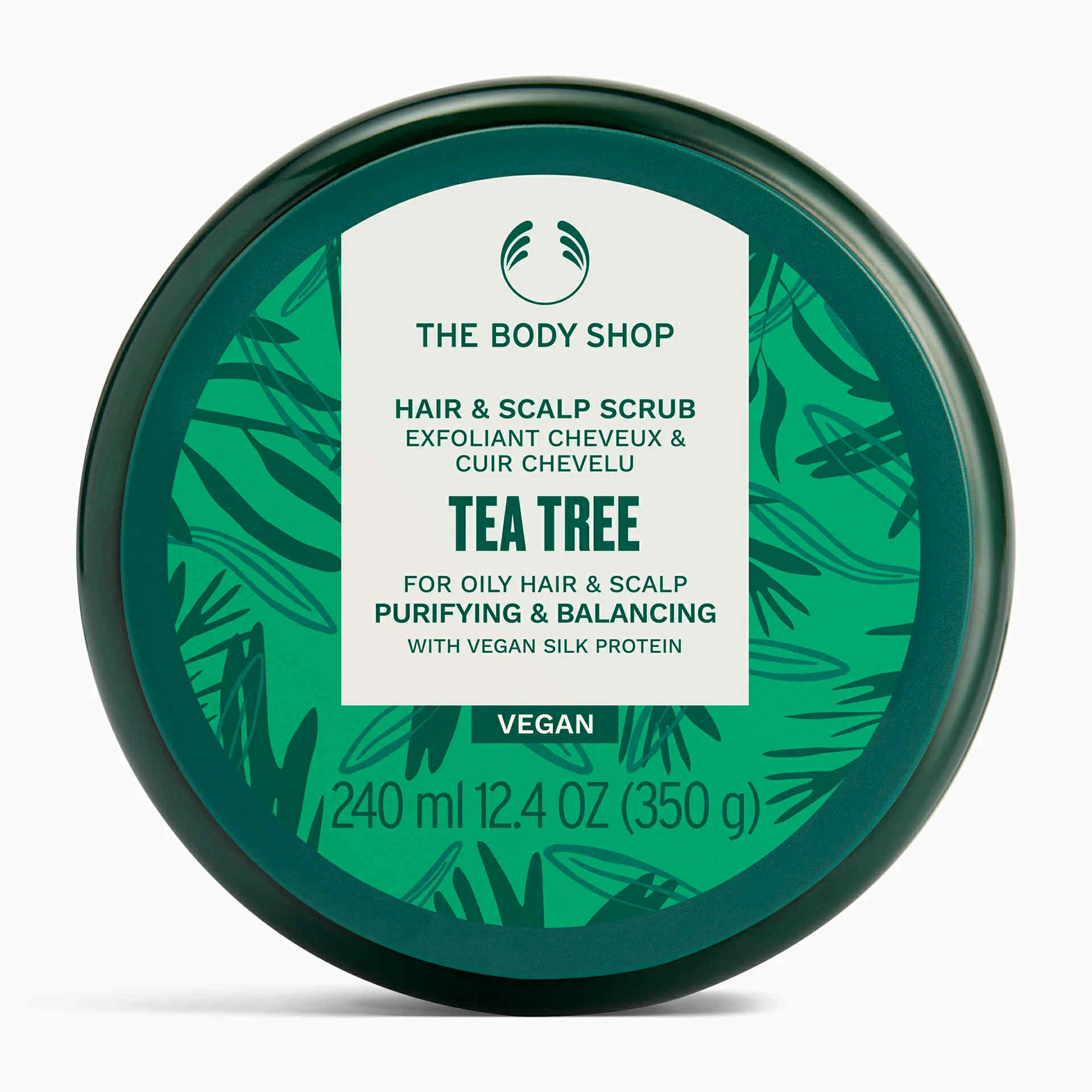 The Scent Of Tea Tree Is So Purifying | The Body Shop Tea Tree Purifying & Balancing Haircare Range A short cylindrical dark green plastic tub with a dark green plastic lid and a bright green label that has The Body Shop Tea tree Purifying & Balancing Hair & Scalp Scrub written in dark green writing and some pictures of some dark green tea tree leaves on it, on a white background.