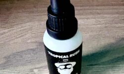 A short cylindrical brown glass bottle with a black rubber pipette lid and a white label that has Thirty 1 Oils Tropical Rush Beard Oil written in bold black writing and a picture of a man wearing a pair of sunglasses on it, on a white background.