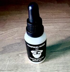 A short cylindrical brown glass bottle with a black rubber pipette lid and a white label that has Thirty 1 Oils Tropical Rush Beard Oil written in bold black writing and a picture of a man wearing a pair of sunglasses on it, on a white background.