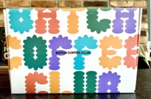A large rectangular bright white cardboard box that has B written in bright teal writing, A written in bright orange writing, t written in dark purple writing, c written in bright green writing, and h written in bright red writing, Batch Coffee Club written in bold black writing, and a picture of a large black oval on it on a dark wooden worktop.