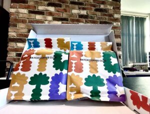 A large rectangular bright white open cardboard box that has two large rectangular bright white Paper Pouches inside it that both have Batch Coffee Club written in bold black writing and some pictures of some bright red, orange, yellow, green, blue and purple wavy lines on them on a dark wooden worktop.