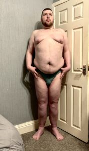 A tall bearded man with his hands on his hips wearing a pair of dark green briefs with a dark green waistband that has Box Menswear written in bold black writing on it stood against a white wooden door.