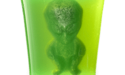 A tall cylindrical translucent lime green block of shower jelly that has a small dark green alien shaped block of shower gummy suspended in the centre of it, on a white background.