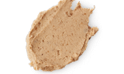 A medium circular puddle of some creamy caramel brown Face Mask that has some pieces of Hazelnut running through it, on a white background.