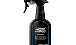 A tall black plastic kitchen spray bottle that has Lush Stormy Weather Body Spray written in bright white writing with a dark blue outline around the words on it, on a white background.