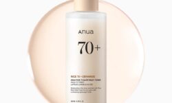 A tall cylindrical frosted white plastic bottle full of some translucent creamy white liquid with a beige plastic lid and a bright white label that has Anua Rice 70 Milky Toner written in bold black writing on it, on a white background.