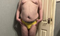 A tall bearded man with his hands on his hips wearing a pair of bright yellow briefs with a black waistband that has Kiniki written in bright white writing on it stood against a white wooden door.