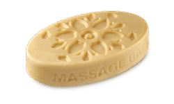 A medium creamy beige oval shaped block of solid massage oil that’s covered in some bright golden yellow lustre and has some pictures of some Flowers and the words Massage Bar engraved on it, on a white background.