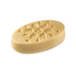 A medium creamy beige oval shaped block of solid massage oil that’s covered in some bright golden yellow lustre and has some pictures of some Flowers and the words Massage Bar engraved on it, on a white background.