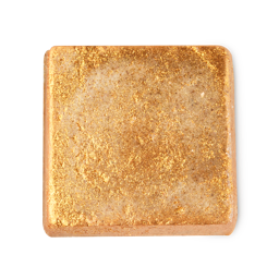 A medium creamy beige square shaped block of solid body scrub that’s coated in some bright gold lustre, on a white background.