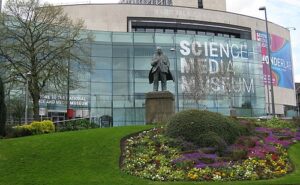 A large rounded rectangular light brown brick building that has some large rectangular glass panes all over it that has National Science and Media Museum written in bright white writing on them next to a tall shiny bronze statue of a man stood on a dark stone pedestal surrounded by some multicoloured flowers against a bright blue sky, on a white background.