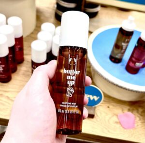 A hand holding a tall cylindrical translucent brown plastic bottle with a creamy white plastic lid and a dark green label that has The Body Shop Butter Me Up Fragrance Mist written in bright white writing and a picture of a bucket full of golden popcorn on it, on a white background.