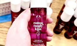 A hand holding a tall cylindrical translucent pink plastic bottle with a creamy white plastic lid and a dark green label that has The Body Shop Cotton Candy Kiss Fragrance Mist written in bright white writing and a picture of some bright pink candy floss on a light wooden stick on it, on a white background.