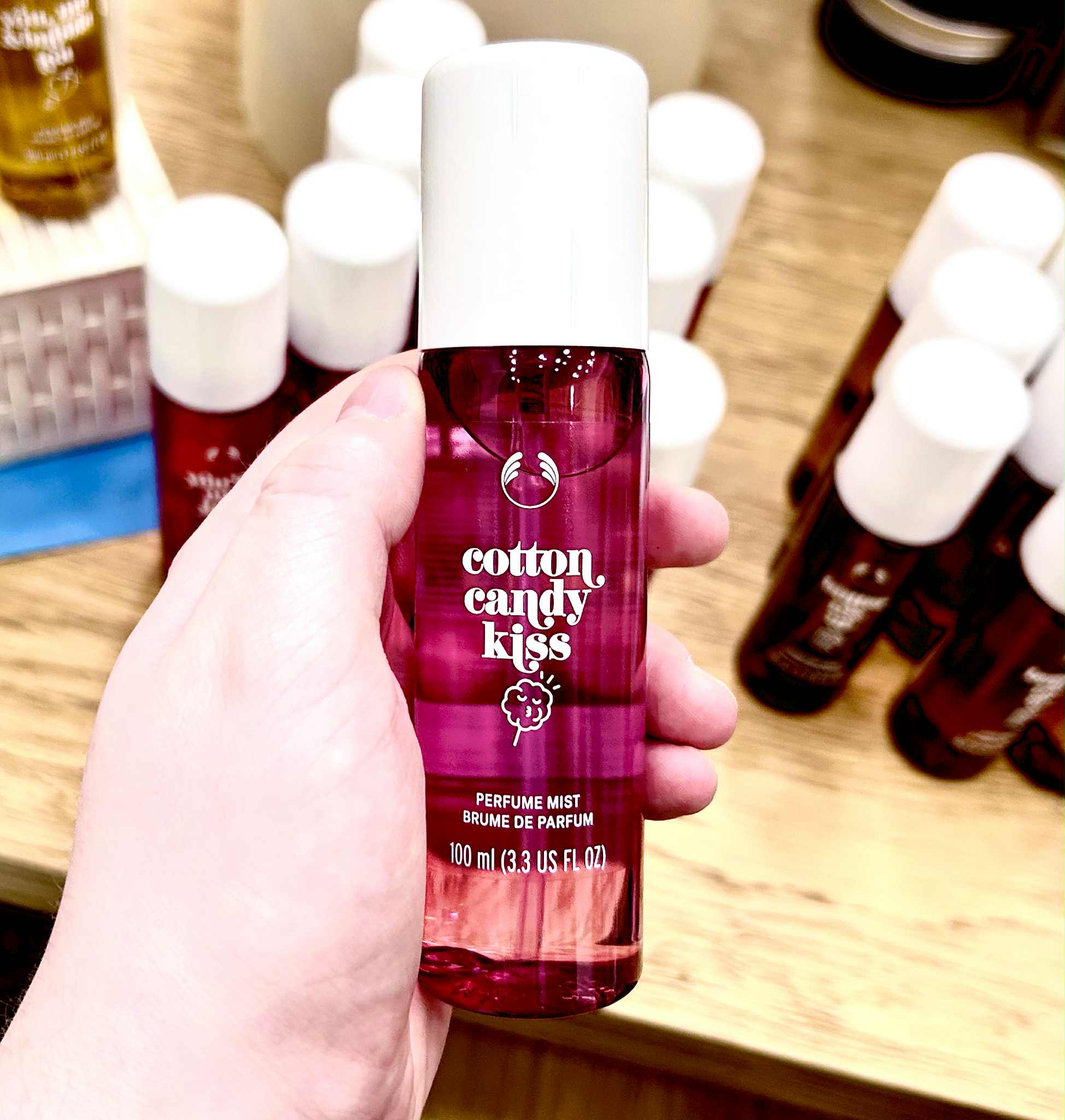 Oh My God! That Body Mist Smells So Sweet | The Body Shop Sweet On You Range A hand holding a tall cylindrical translucent pink plastic bottle with a creamy white plastic lid and a dark green label that has The Body Shop Cotton Candy Kiss Fragrance Mist written in bright white writing and a picture of some bright pink candy floss on a light wooden stick on it, on a white background.