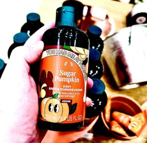 A hand holding a tall cylindrical dark green plastic Bottle with a dark green plastic lid and a bright orange label that has The Body Shop Sugar Pumpkin Bath & Shower Cream written in bright white writing and some pictures of some large bright orange pumpkins with some dark green leaves on it, on a white background.