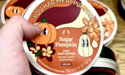 A hand holding a short cylindrical dark green plastic tub with a dark green plastic lid and a bright orange label that has The Body Shop Sugar Pumpkin Body Butter written in bright white writing and some pictures of some bright orange pumpkins with some dark green leaves on it, on a white background.