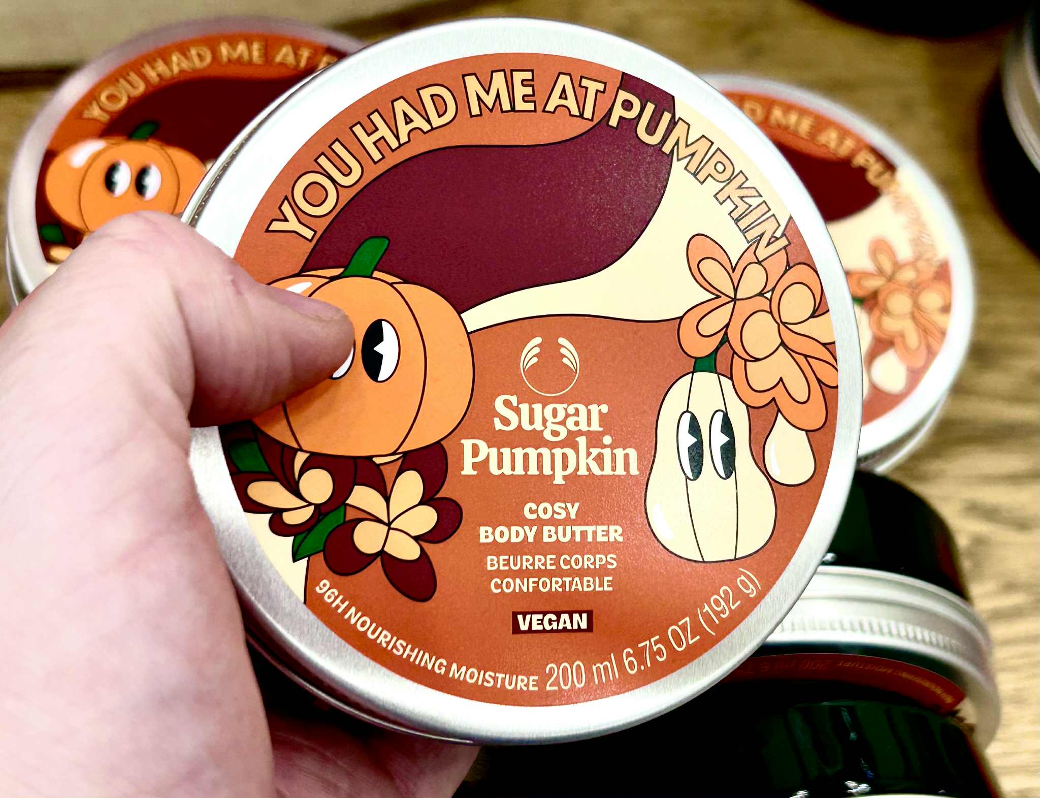 Sweet!! It's Finally Pumpkin Season | The Body Shop Sugar Pumpkin Range A hand holding a short cylindrical dark green plastic tub with a dark green plastic lid and a bright orange label that has The Body Shop Sugar Pumpkin Body Butter written in bright white writing and some pictures of some bright orange pumpkins with some dark green leaves on it, on a white background.