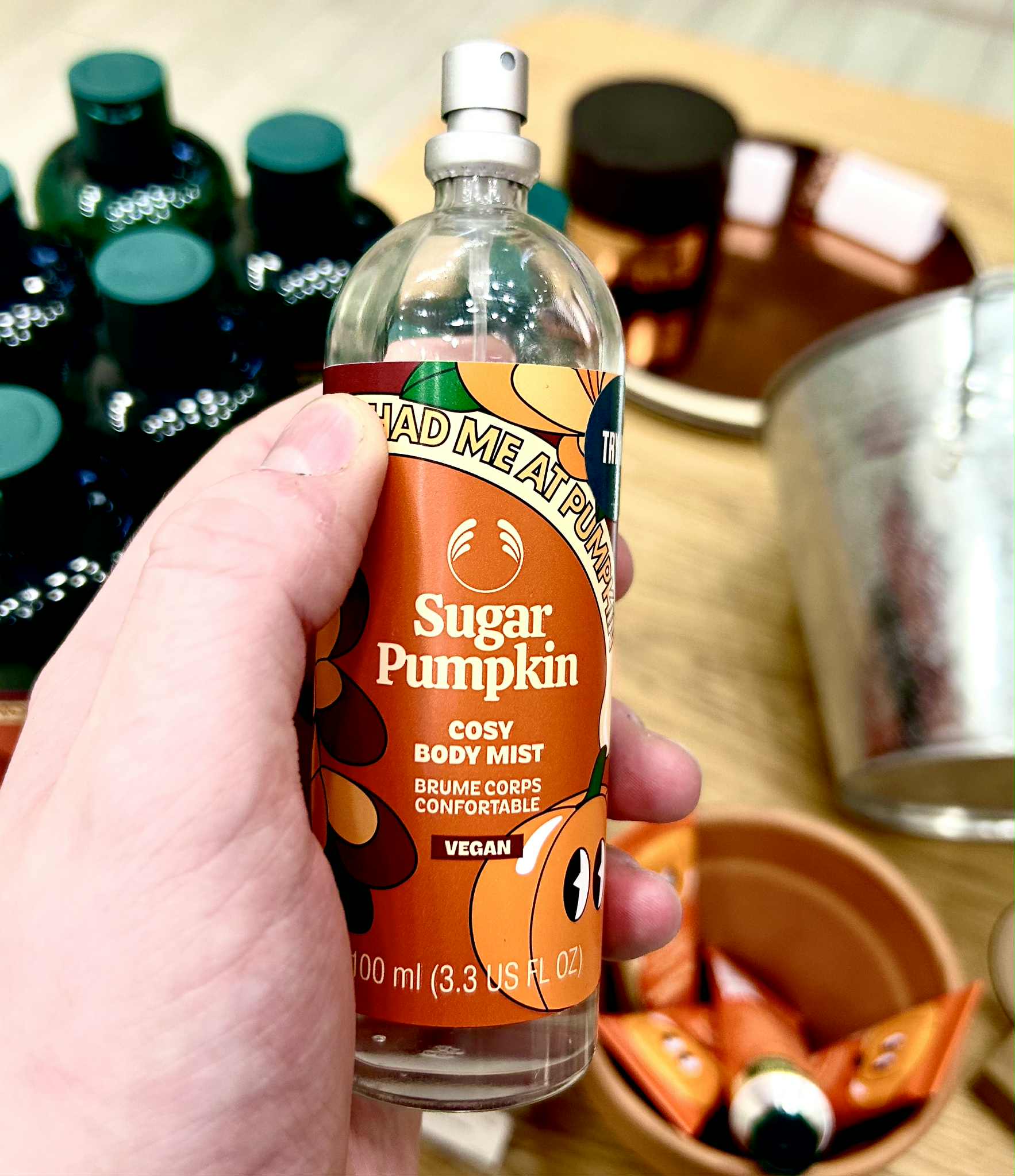 A tall cylindrical clear glass bottle full of some clear liquid with a silver metal lid and a bright orange label that has The Body Shop Sugar Pumpkin Body Mist written in bright white writing and a picture of some bright orange pumpkins with some dark green leaves on it, on a white background.