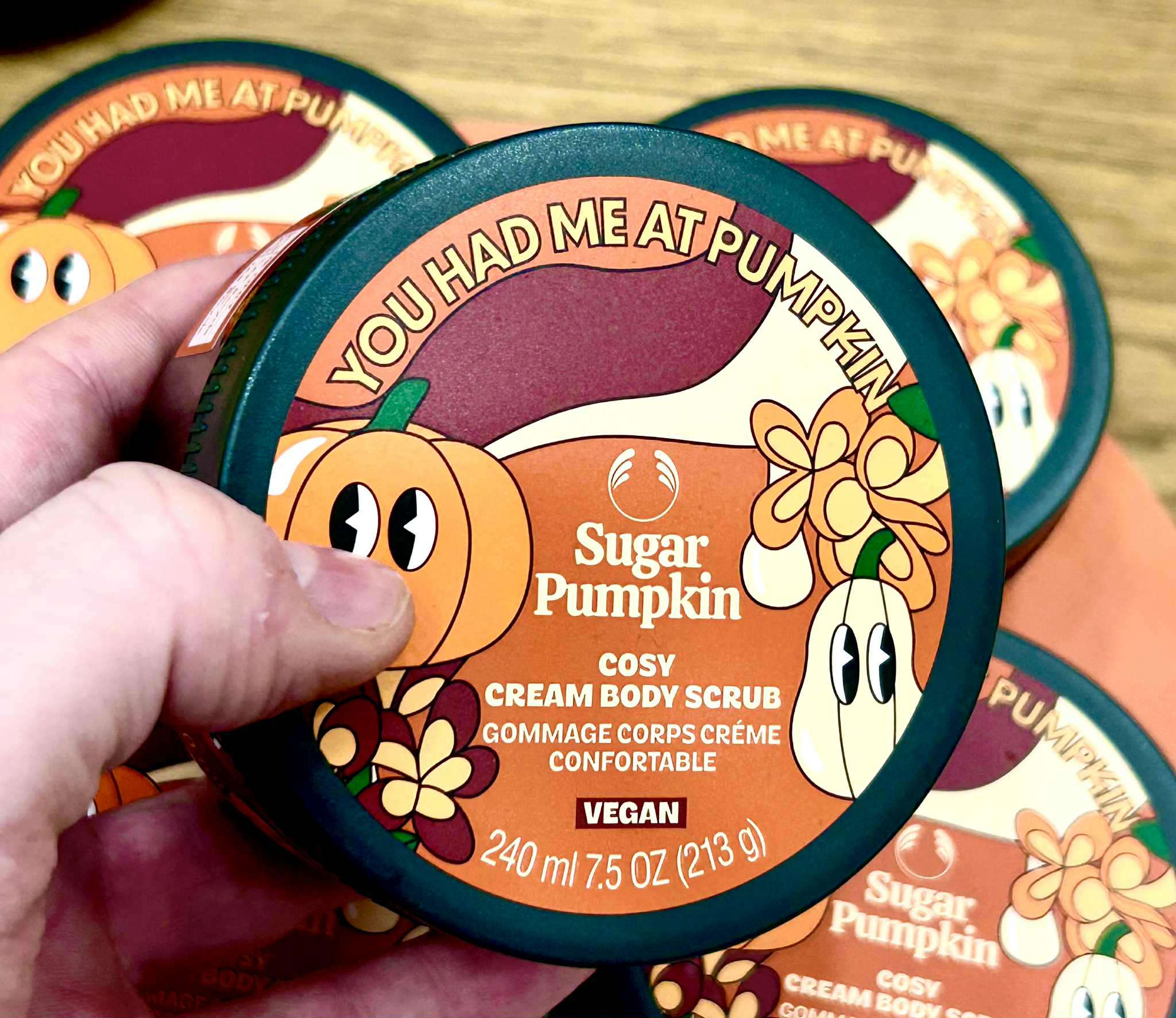 A hand holding a short cylindrical dark green plastic tub with a dark green plastic lid and a bright orange label that has The Body Shop Sugar Pumpkin Body Scrub written in bright white writing and some pictures of some bright orange pumpkins with some dark green leaves on it, on a white background.