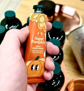 A hand holding a long and thin conical metallic bright orange tube with a metallic dark green lid and a bright orange lable that has The Body Shop Sugar Pumpkin Hand Balm written in bright white writing and some pictures of some bright orange pumpkins with some dark green leaves on it, on a white background.