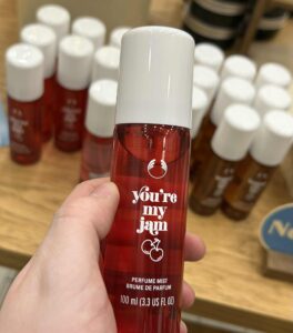 A hand holding a tall cylindrical translucent red plastic bottle with a creamy white plastic lid and a dark green label that has The Body Shop You’re My Jam Fragrance Mist written in bright white writing and a picture of some bright red cherries on it, on a white background.