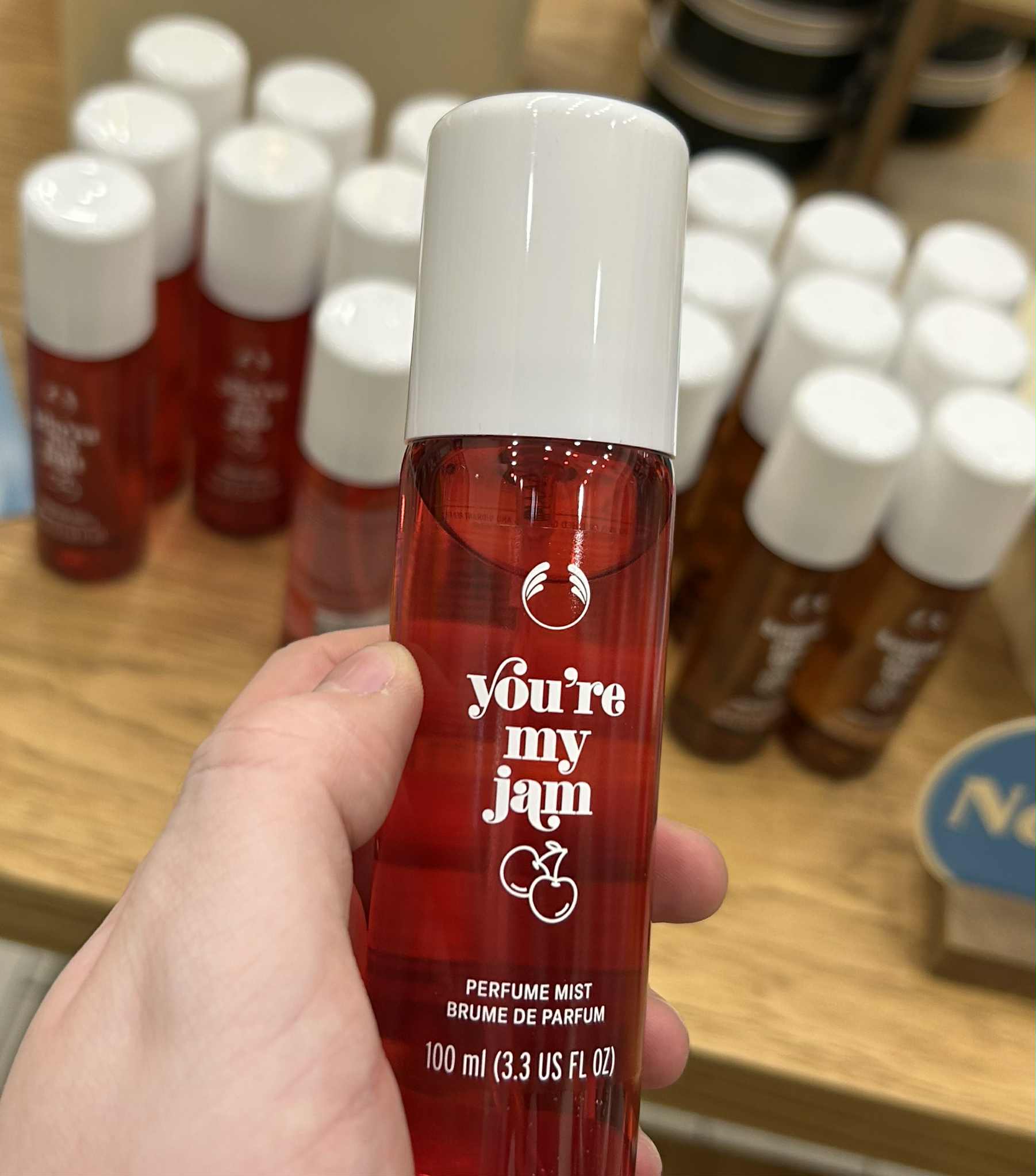 A hand holding a tall cylindrical translucent red plastic bottle with a creamy white plastic lid and a dark green label that has The Body Shop You’re My Jam Fragrance Mist written in bright white writing and a picture of some bright red cherries on it, on a white background.