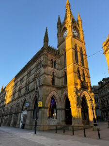 A tall historic gothic light brown stone building and Clock tower that has some pointed arches and carvings all over it next to a light brown stone paved street that has some silver metal lamp posts and some string lights dotted all around it against a light blue sky, on a white background.