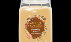 A tall cylindrical glass jar full of some pale brown wax with a label that has Yankee Candle written in bright white writing, glistening Leaves written in bright white writing, and a picture of some golden orange leaves, some oranges, a slice of Fig, and a gold ribbon on it, on a black background.