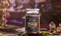 A tall cylindrical glass jar full of some dark purple wax with a label that has Yankee Candle written in white writing, Mystical Tarot Card written in white writing, and a picture of a small rectangular pale purple Tarot Card with a black spiral design in the centre of it next to a small rectangular pale blue Tarot Card with two dark red cherries in the centre of it, and a small rectangular pale orange tarot card that has two coiled snakes in the centre of it on it, on a white background.