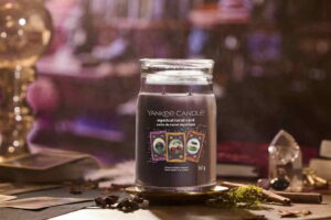 A tall cylindrical glass jar full of some dark purple wax with a label that has Yankee Candle written in white writing, Mystical Tarot Card written in white writing, and a picture of a small rectangular pale purple Tarot Card with a black spiral design in the centre of it next to a small rectangular pale blue Tarot Card with two dark red cherries in the centre of it, and a small rectangular pale orange tarot card that has two coiled snakes in the centre of it on it, on a white background.